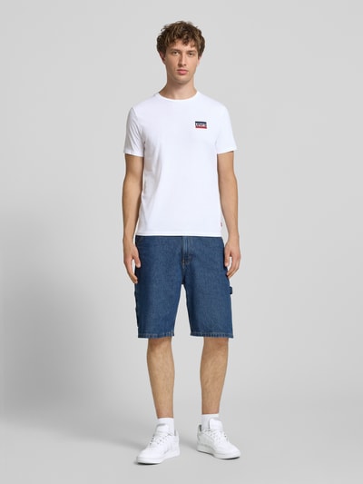 Männermodel trägt weißes Slim Fit T-Shirt-Set aus reiner Baumwolle im 2er-Pack von Levi’s®, kombiniert mit blauer Jeansshorts, Ganzkörperansicht von vorne.