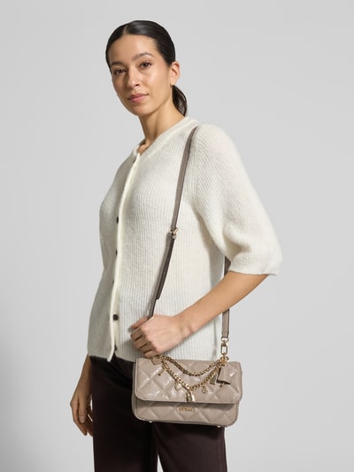 Frauenmodel trägt Umhängetasche mit Label-Applikation Modell 'LIBBY' von Guess in Beige mit Ketten-Detail, Nahaufnahme Oberkörper.