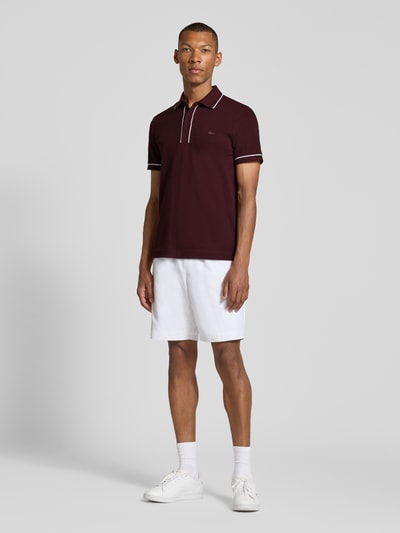 Mannenmodel draagt Regular fit poloshirt uit katoenmix van Lacoste in donkerrood met witte details, witte korte broek, sneakers, geheellichaamsaanzicht van voren.