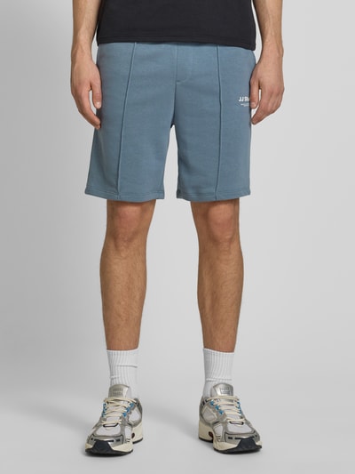 Męskamodel nosi Sweatshorts mit Logo Model 'KANE' od Jack & Jones, bawełniany miks, krój relaxed fit, elastyczny pas, logo, szerokość w rozmiarze M: 42 cm, długość wewnętrzna nogawki w rozmiarze M: 19 cm, kolor niebieski, tekstura gładka, widok całej sylwetki z przodu.