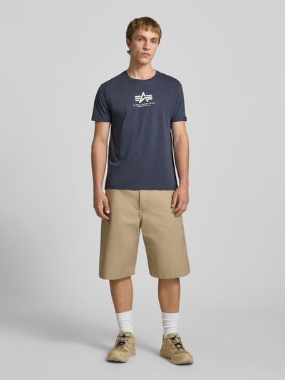 Mannelijk model draagt T-Shirt met Logo en Rundhalsausschnitt van Alpha Industries, regular fit, katoen, logo, marineblauw, voorkant volledig zichtbaar, Ganzkörperansicht von vorne.