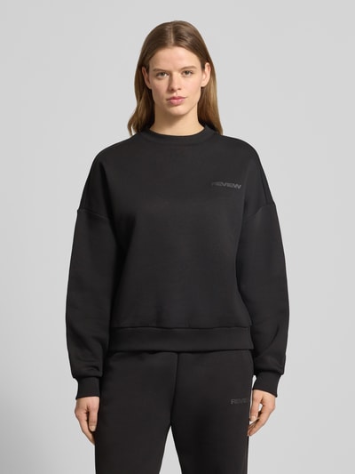 Frauenmodel trägt schwarzen Basic Sweater mit Rundhalsausschnitt und Bündchen am Ärmel von Review, Oversized, Label-Print, Logo-Stickerei, Ganzkörperansicht von vorne.