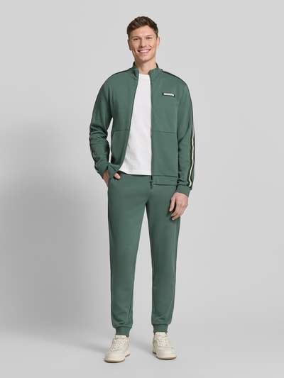 Männermodel trägt Sweatjacke mit Stehkragen und Zweiwege-Reißverschluss von EA7 Emporio Armani in Grün mit Label-Detail, Ganzkörperansicht von vorne.