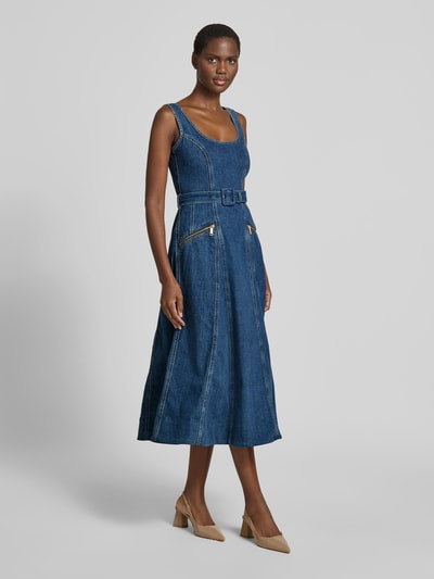Frauenmodel trägt Mini-Jeanskleid aus Baumwoll-Mix von Self-Portrait, Midikleid in Denim-Optik, taillierte Passform, A-Linie, ärmelloses Design, Farbtöne in Dunkelblau mit goldfarbenen Zippern und Gürtel, strukturierte Textur, Ganzkörperansicht von vorne.