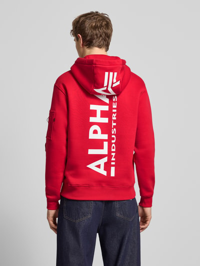 Männermodel trägt roten Hoodie mit Känguru-Tasche von Alpha Industries aus Baumwoll-Mix, Regular-Fit, Label-Patch, Ganzkörperansicht Rückansicht.
