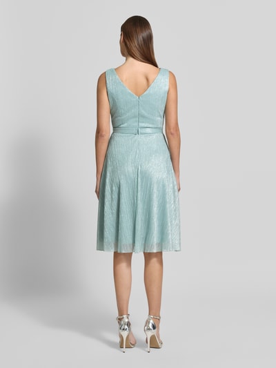 Frauenmodel trägt knielanges Cocktailkleid mit V-Ausschnitt von TROYDEN COLLECTION, schimmernd, taillierter Schnitt, Taillenband, Ganzkörperansicht Rückansicht.
