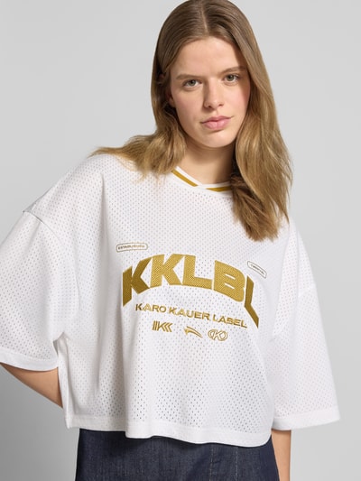 Frauenmodel trägt Cropped T-Shirt mit rückseitigem Logo-Print von Karo Kauer in Weiß mit Label-Print, Loose-Fit, Rundhalsausschnitt, Label-Stitching, Rückenlänge 47 cm, Nahaufnahme Oberkörper.