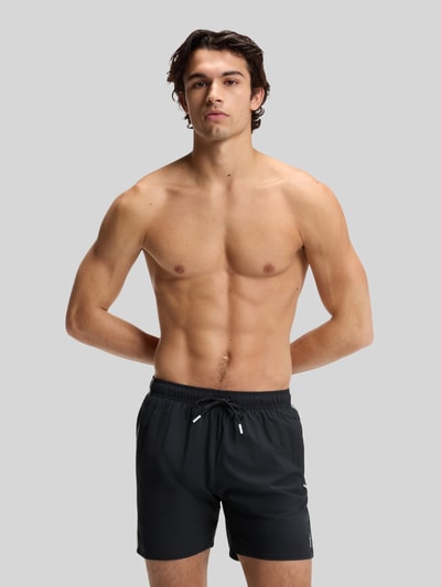 Männermodel trägt Badeshorts mit Tunnelzug Modell 'ICONIC' von BOSS, unifarben mit Label Print und Kontraststreifen, Ganzkörperansicht von vorne.