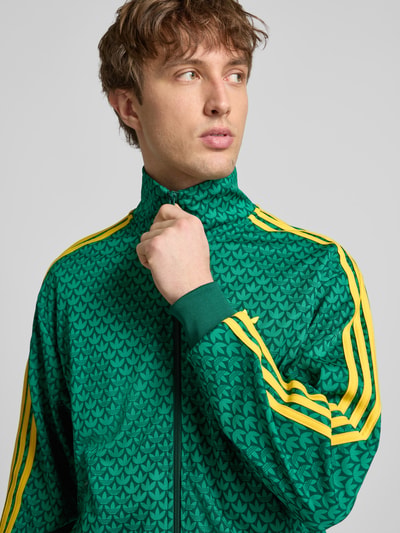 Männermodel trägt Slim Fit Jacke mit Stehkragen Modell 'Firebird' von adidas Originals in Grün mit gelben Streifen und Logo-Stitching, Nahaufnahme Oberkörper.
