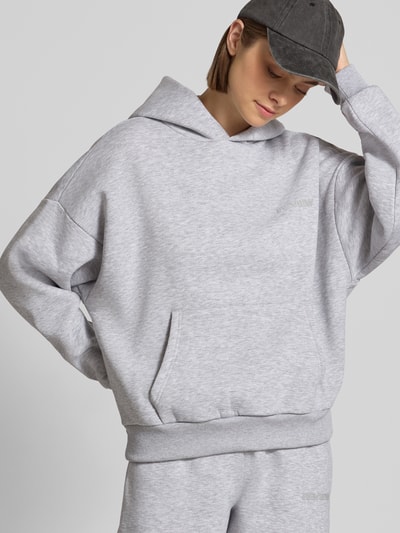 Kobiece model prezentuje Relaxed Fit Basic Hoodie in grau melange od REVIEW, mieszanka bawełny, kaptur, nadruk z logo, kieszeń kangurka, prążkowane wykończenia, widok na górną część ciała z przodu.