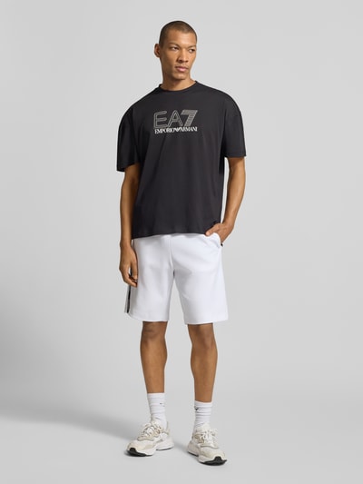 Męski model trägt T-shirt mit Logo und Rundhalsausschnitt von EA7 Emporio Armani, bawełna, krój regular fit, długość tyłu 68 cm, Ganzkörperansicht von vorne.