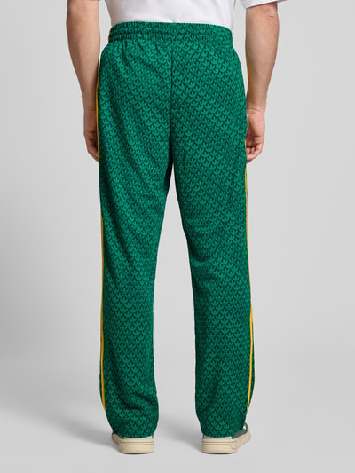 Männermodel trägt grüne Sweatpants mit elastischem Bund von adidas Originals, lockerer Schnitt, gelbe Seitenstreifen, Ganzkörperansicht Rückansicht.