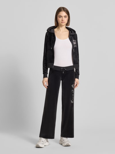 Modelka nosi czarne Sweatpants mit Ziersteinbesatz Modell 'GOTHIC CROWN' od Juicy Couture, materiał elastyczny, krój straight fit, ozdobny sznurek i elastyczny pas; damska moda, widok całej sylwetki z przodu.