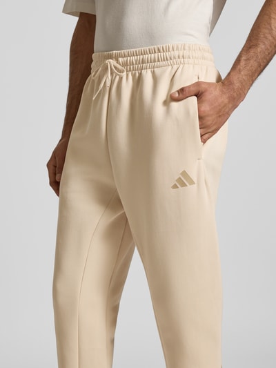 Männermodel trägt Regular Fit Sweathose mit Logo-Print van adidas Sportswear in beige katoenmix met elastische band en tunnelkoord, logoprint, elastische beenboorden, binnenbeenlengte maat M 71 cm, nahaufnahme heup- en bovenbeen.