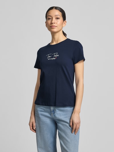 Modelka nosi Regular Fit T-Shirt aus Bio-Baumwoll-Mix von Tom Tailor, krój regular fit, jednolity kolor, krótki rękaw, okrągły dekolt, aplikacja z napisem, materiał: mieszanka bio-bawełny, widok całej sylwetki z przodu.