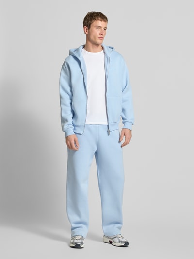 Männermodel trägt Regular Fit Sweatjacke mit Reißverschluss von Review aus hellblauem Baumwoll-Mix, Logo-Print, Kapuze, Kängurutasche, elastische Abschlüsse; Ganzkörperansicht von vorne.