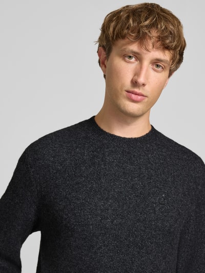 Männermodel trägt Strickpullover mit Logo-Stitching Modell 'CURTIS' von Carhartt Work In Progress, meliertes Dunkelgrau, Nahaufnahme Oberkörper. Material-Mix mit Stretch-Anteil, lockerer Schnitt, Rundhalsausschnitt, gerippte Abschlüsse.