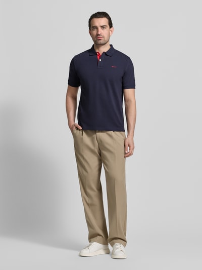 Męski model nosi Regular Fit Poloshirt mit Logo-Stitching od Gant, granatowy, bawełniany, z kołnierzykiem polo, krótkim rękawem, krój regular fit, widok całej sylwetki z przodu.