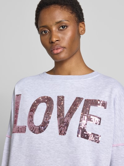 Vrouwenmodel draagt oversized dames sweatshirt van puur katoen Model 'Love' van miss goodlife met ronde hals, statementprint met pailletten, elastische boorden. Nahaufnahme bovenlichaam.