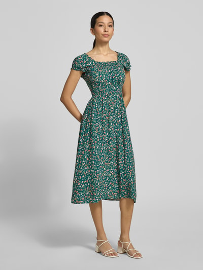 Kobiece modelka nosi Midikleid aus Viskose mit Smok-Details od Apricot, wzór na całej powierzchni, taliowany fason, długość tyłu w rozmiarze S: 110 cm, szerokość w rozmiarze S: 40 cm, zielone tło z kwiatowym wzorem, lekka tekstura, całkowity widok z przodu.