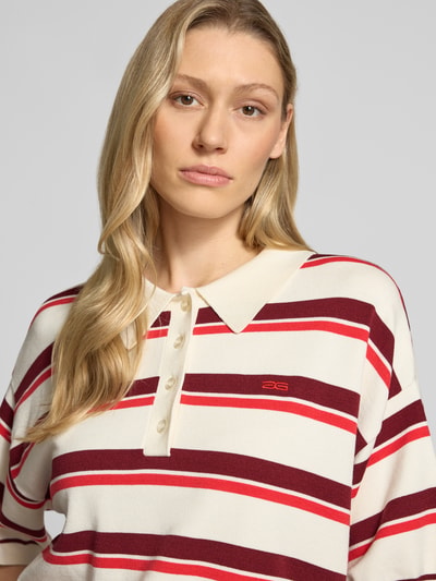 Modelka nosi koszulkę polo, mieszanka wiskozy, krój relaxed fit, model 'Taia' od Gestuz, imitacja dzianiny, rękaw o dł. 1/2, elastyczne wykończenia, długość tyłu w rozmiarze S: 58 cm, długość rękawów w rozmiarze S: 22 cm, paski w odcieniach kremowym, bordowym i czerwonym, widoczna faktura materiału, zbliżenie na górną część ciała, zdjęcie z przodu