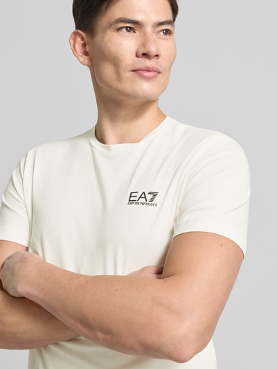 Mannelijk model draagt T-Shirt met Label-Print van EA7 Emporio Armani in crème, slim fit, katoenen mix, Nahaufnahme bovenlichaam.
