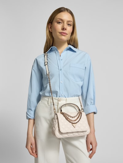Frauenmodel trägt Handtasche mit Steppnähten Modell 'GIULLY II' von Guess, Überschlag, Magnetverschluss, Label-Applikation, Tragehenkel, Schulterriemen, Hauptfach, Breite 21 cm, Länge 16 cm, Tiefe 6 cm, Länge Schulterriemen 116 cm, helle Farben, glatte und gesteppte Textur, Ganzkörperansicht von vorne.