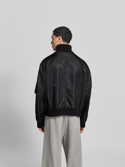 Männermodel trägt Regular Fit Bomberjacke mit Stehkragen von MM6 Maison Margiela in Schwarz, elastische Abschlüsse, Eingrifftaschen, Ärmeltasche, Reißverschluss, Rückenlänge 63 cm, Schulterbreite 54 cm, Ärmellänge 67 cm, Ganzkörperansicht Rückansicht.