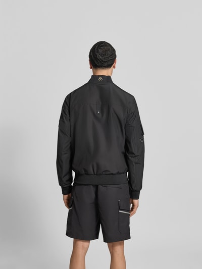 Männermodel trägt schwarze Bomberjacke mit Zweiwege-Reißverschluss von Moose Knuckles, lockere Passform, Rückenansicht, elastische Abschlüsse, Stehkragen, Reißverschluss- und Ärmeltaschen, Rückenlänge 67 cm, Schulterbreite 46 cm, Ärmellänge 68 cm, Ganzkörperansicht Rückansicht.