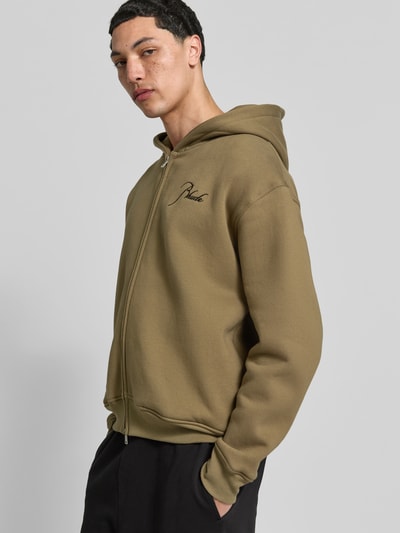 Männermodel trägt Oversized Sweatjacke aus Baumwolle von RHUDE in Oliv mit Zweiwege-Reißverschluss und Label-Stitching, Nahaufnahme Oberkörper.