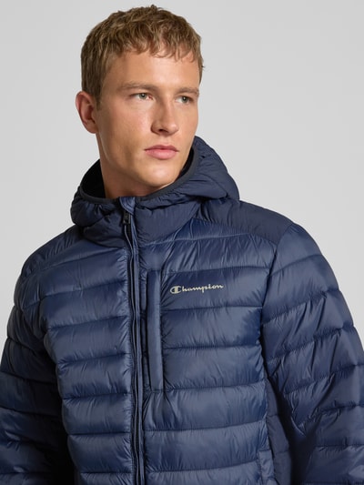 Mannelijk model draagt Steppjacke met Kapuze und Logo van CHAMPION in regular fit, donkerblauw, close-up bovenlichaam, zicht op logo.