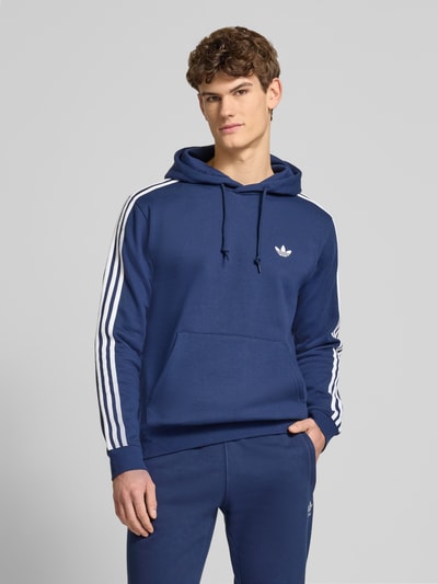 Męski model nosi Hoodie mit Kapuze od adidas Originals w niebieskim kolorze, bawełniany miks, krój luźny, logo, kieszeń kangurka, prążkowane wykończenia, długość tyłu w rozmiarze M 70 cm, długość rękawa w rozmiarze M 67 cm, widok całej sylwetki z przodu.