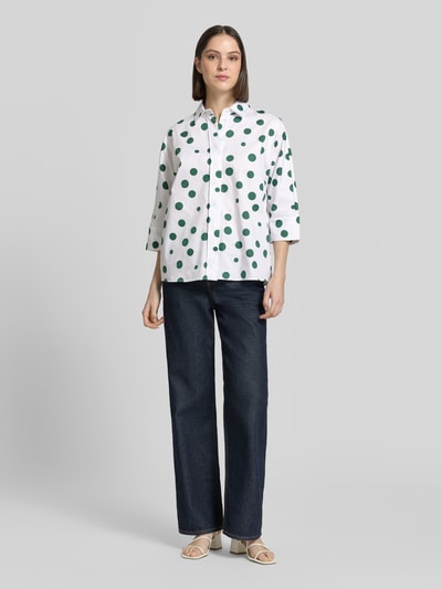 Frauenmodel trägt Relaxed Fit Hemdbluse mit 3/4-Arm von Christian Berg Woman, weiße Reine Baumwolle mit grünen Punkten und durchgehender Knopfleiste, Hemdblusenkragen, kombiniert mit dunkler Hose, Ganzkörperansicht von vorne.