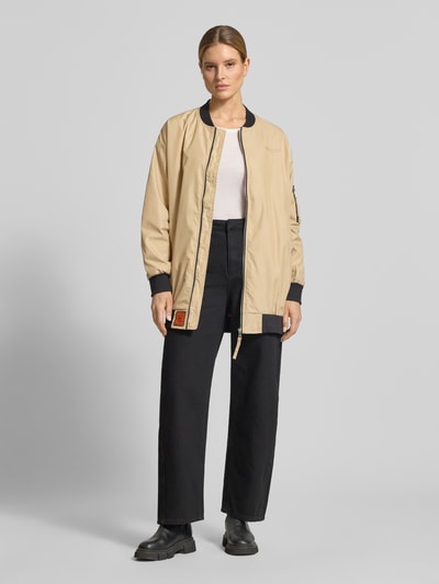 Frauenmodel trägt Relaxed Fit Jacke mit Logo-Stitching von Original Bombers in Beige mit Stehkragen, Reißverschluss, elastischen Abschlüssen, Eingrifftaschen und Armtasche; Jacke wirkt glatt, Kombi mit hellen und dunklen Farben, Ganzkörperansicht von vorne.
