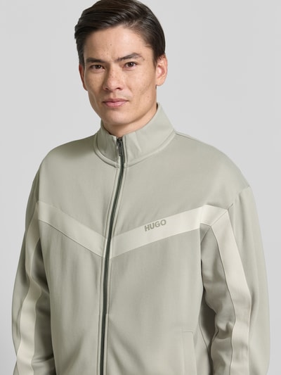 Mężczyznamodel nosi Relaxed Fit Sweatjacke mit Baumwoll-Anteil Modell 'DARTI' od HUGO, gładki materiał, zamek, detal z logo, taśma w jodełkę, stojący kołnierz, długie rękawy, boczne kieszenie, kolor jasnoszary, zbliżenie na górną część ciała.