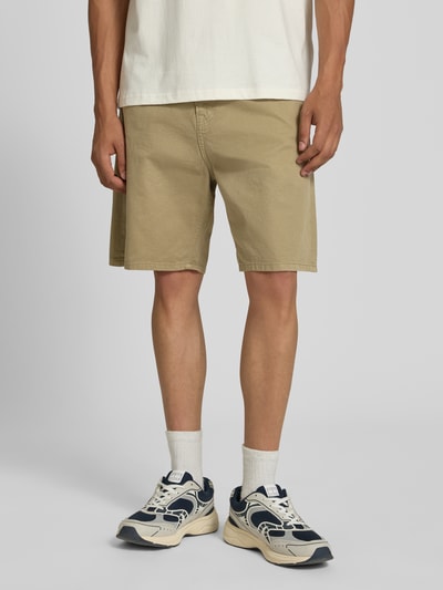 Męski model nosi bermudy z bawełny w kolorze beżowym, 5 kieszeni, krój straight fit, model 'TONY ORIGINAL' od Jack & Jones, szerokość w rozmiarze M: 42 cm, długość wewnętrzna nogawki w rozmiarze M: 20 cm, zapięcie na guzik i zamek błyskawiczny, Ganzkörperansicht von vorne.