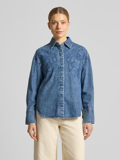 Modelka nosi Loose Fit Jeansbluse aus reiner Baumwolle od s.Oliver RED LABEL w kolorze niebieskiego denimu, luźny krój, haft z motywem na piersi, kołnierzyk koszulowy, długi rękaw z mankietem, widoczna faktura twillu, bawełna, widok całej sylwetki z przodu.
