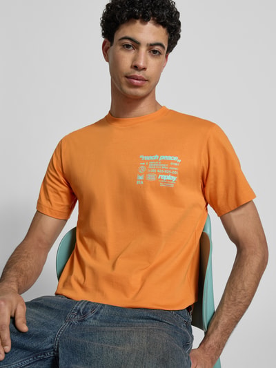 Męski model nosi Regular Fit T-Shirt mit Statement-Print od Replay, czysta bawełna, krój regular fit, okrągły dekolt, długość pleców w rozmiarze M: 62 cm, kolor pomarańczowy, nadruk, tekstura gładka. Ujęcie: zbliżenie na górną część ciała z przodu.