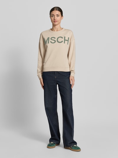 Modelka nosi bluzę Sweatshirt mit Label-Stitching Modell 'Mela Briena' od MSCH Copenhagen z mieszanki bawełny, krój loose fit, okrągły dekolt, wyhaftowane logo, jasny beż z zielonym nadrukiem, pełen widok z przodu.