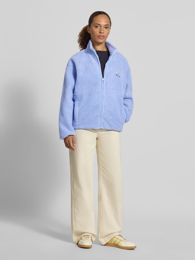 Frauenmodel trägt Regular Fit Jacke aus Teddyfell Modell 'Hoktown' von American Vintage in Hellblau mit Stehkragen, Logo-Patch und Reißverschluss, Eingrifftaschen, Struktur und Schnitt aus dem Kontext ersichtlich, Ganzkörperansicht von vorne.