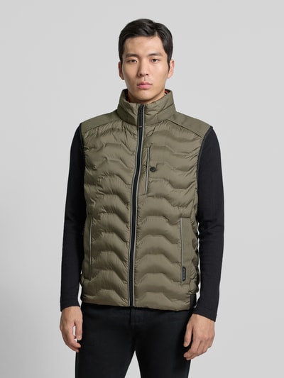 Męski model nosi Regular Fit Steppweste mit Kapuze im Kragen od Tom Tailor, kolor jednolity, krój regular fit, stójka z kapturem w kołnierzu, zamek błyskawiczny, label patch, odblaskowe detale, boczne kieszenie na zamek, kieszeń wewnętrzna, widok całej sylwetki z przodu.