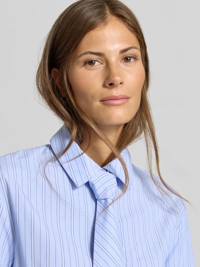 Frauenmodel trägt Hemdbluse aus reiner Baumwolle mit integrierter Krawatte von MYON, hellblau mit feinen Streifen, Nahaufnahme Oberkörper. Taillierte Passform, Hemdblusenkragen, langärmeliges Design, Rückenlänge 58 cm, Ärmellänge 64 cm.