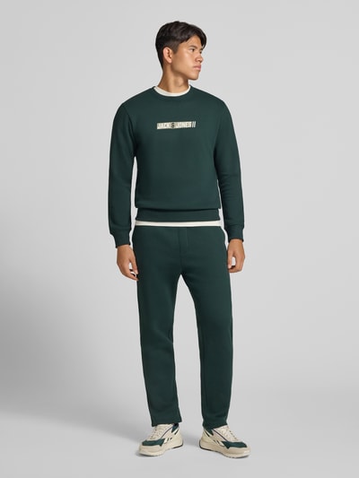 Männermodel trägt Regular Fit Sweatshirt mit Baumwoll-Anteil Modell 'Munk' von JACK & JONES in Dunkelgrün mit Logo-Print, Ganzkörperansicht von vorne. Material-Mix, elastische Abschlüsse, Rundhalsausschnitt.