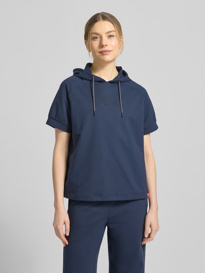 Modelka kobieca nosi T-Shirt aus Baumwoll-Mix mit Kapuze Modell 'SHARON3' od FIRE + ICE, luźny krój, granatowy, krótki rękaw, kaptur ze ściągaczem, elastyczne wykończenie rękawów, detal z logo, widok całej sylwetki z przodu.