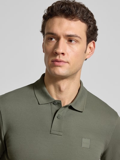 Männermodel trägt Slim Fit Poloshirt aus Baumwoll-Mix Modell 'PASSENGER' von BOSS Orange in Olivgrün, Label Patch, Nahaufnahme Oberkörper.