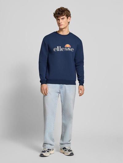 Männermodel trägt Sweatshirt mit Label-Print Modell 'CORVARO' von ellesse in Dunkelblau, Material-Mix mit Viskose-Anteil, lockerer Schnitt, gerippte Abschlüsse, Rundhalsausschnitt, Rückenlänge 69 cm, Ärmellänge 64 cm, Ganzkörperansicht von vorne.