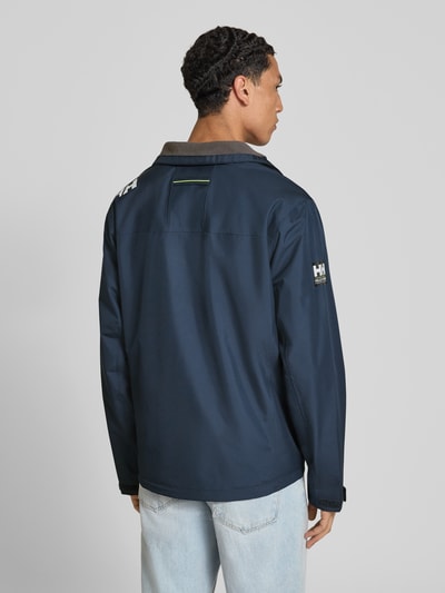 Männermodel trägt Regular Fit Jacke mit Stehkragen Modell 'Crew' von Helly Hansen in Dunkelblau, glatte Oberfläche, Ganzkörperansicht Rückansicht. Herren Jacke mit Logo-Print, Napoleontasche, Reißverschlusstaschen, Länge 73 cm, Schulterbreite 48 cm, Brustweite 56 cm, Ärmellänge 68 cm.