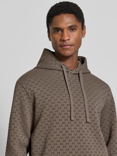 Männermodel trägt Regular Fit Hoodie aus Baumwoll-Mix von Armani Exchange in Braun mit Allover-Logo-Print, Nahaufnahme Oberkörper. Rückenlänge 66 cm, Ärmellänge 63 cm, Kapuze mit Tunnelzug, elastische Abschlüsse.
