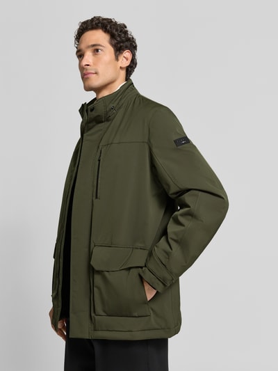 Mężczyzna nosi Jacke mit Pattentaschen und Ärmelriegeln od bugatti, ciemnozielony kolor, krój regular fit, Kaptur, stójka, zamek błyskawiczny, Kieszeń zapinana na zamek błyskawiczny, kieszenie z patką, Patki przy mankietach, Długość tyłu w rozmiarze 50: 80 cm, Szerokość ramion w rozmiarze 50: 50 cm, Szerokość w rozmiarze 50: 62 cm, Długość rękawów w rozmiarze 50: 67 cm, Ganzkörperansicht z przodu.