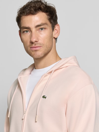 Männermodel trägt Regular Fit Sweatjacke aus Baumwoll-Mix von Lacoste in zartem Rosa, glatte Struktur, Nahaufnahme Oberkörper.